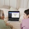 A joyful woman and her grandma browse a beautifully designed GED diploma template on a laptop while in bed, highlighting the value of a well-presented diploma in job applications.