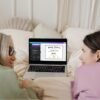 Sharing smiles and success: A happy granddaughter and her grandma together in bed, with a laptop displaying a GED graduation certificate template on Canva.